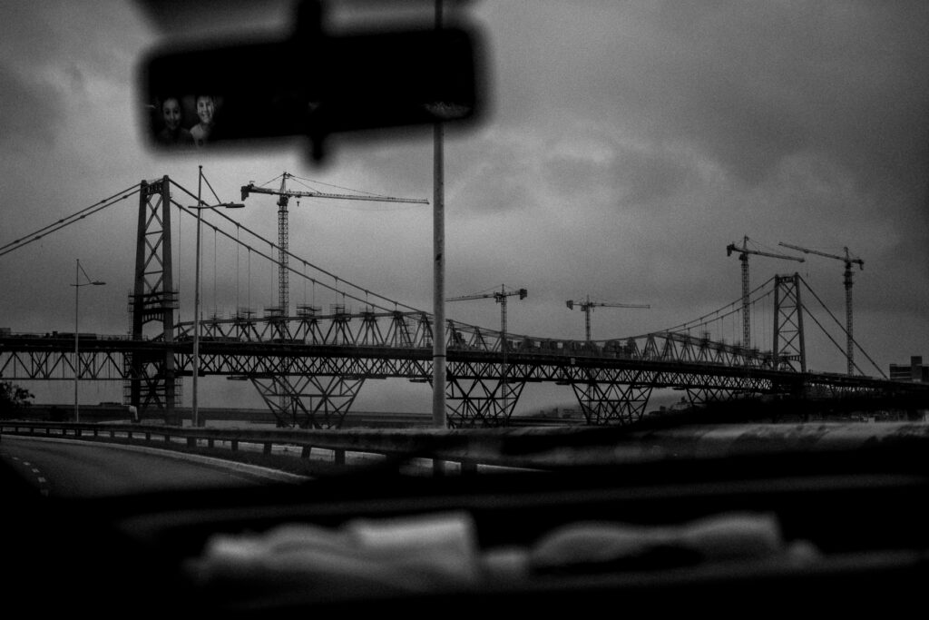 Fotografia em preto e branco feita da janela do carro. No plano geral, a ponte Hercílio Luz de Florianópolis está no horizonte, cobrindo a paisagem. No canto superior esquerdo, levemente centralizado, o retrovisor do carro reflete duas crianças creepy sorrindo. A imagem foi capturada ao acaso; as crianças são reflexo de um outdoor.